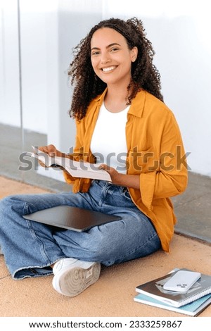 Vertical photo of a lovely stylish curly haired brazilian or hispanic female student, sit near the university campus, reading a book, preparing for the exam, doing homework, looks and smile at camera