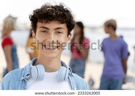 Portrait of smiling teenager with braces wearing headphones looking at camera standing on the street with friends on background. Summer concept Royalty-Free Stock Photo #2333262005
