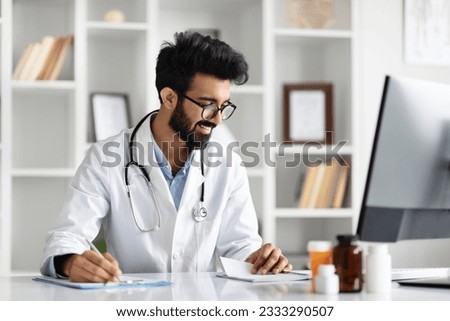 Handsome indian man doctor using computer and writing notes in medical journal sitting at desk. Young professional medic physician wearing eyeglasses, white coat and stethoscope working at clinic