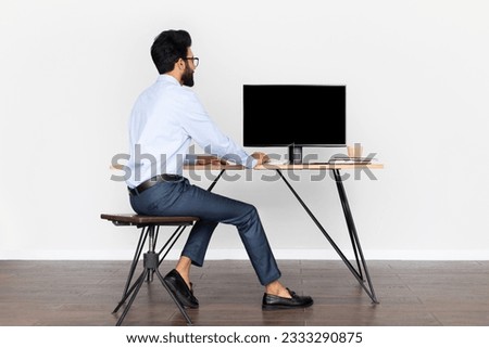 Back view of handsome young arab businessman looking at pc computer with blank black screen for mockup. Eastern man sitting at desk at home office, free space. People, technology, business concept Royalty-Free Stock Photo #2333290875