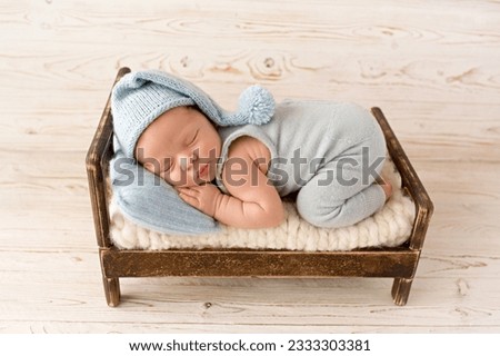 A cute newborn boy in the first days of life sleeps in a blue overalls on a wooden brown bed. Blue knitted hat and Blue head pillow. Against a light wooden background. Royalty-Free Stock Photo #2333303381