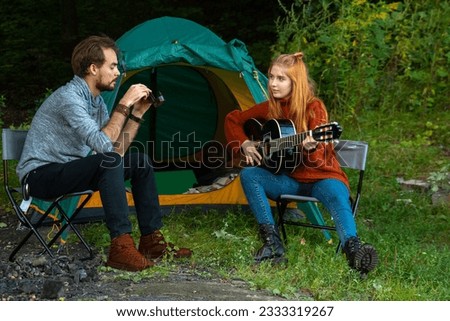 Young man taking photos of girlfriend playing guitar while spending time at campsite together. One shot in series of camping 