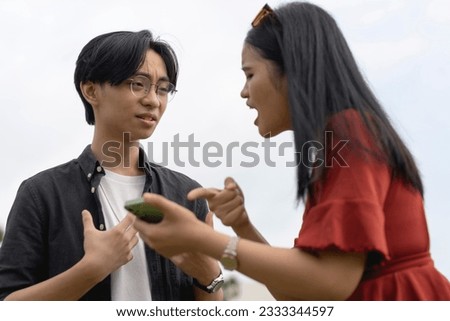 A young asian woman confronts her cheating boyfriend over a extramarital relationship with another girl on his phone. A man taken aback and surprised after getting caught red-handed. Royalty-Free Stock Photo #2333344597