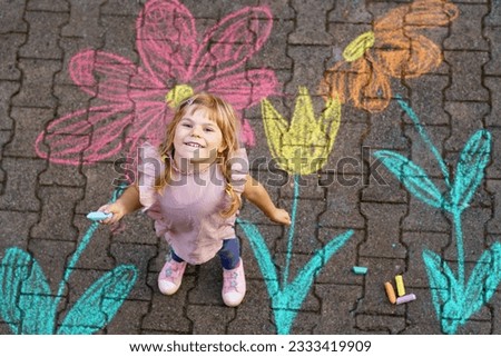 Little preschool girl painting with colorful chalks flowers on ground on backyard. Positive happy toddler child drawing and creating pictures on asphalt. Flower for mother's day