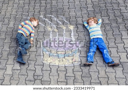 Two happy little kids having fun with big birthday cake picture drawing with colorful chalks. Creative leisure for children outdoors in summer. Kids blowing candles