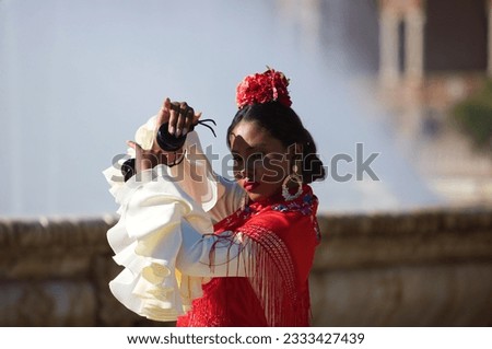 Young black woman dressed as a flamenco gypsy in a famous square in Seville, Spain. She is wearing a beige dress with ruffles and red shawl and playing castanets.Flamenco cultural heritage of humanity Royalty-Free Stock Photo #2333427439