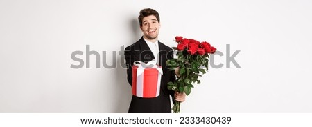 Concept of holidays, relationship and celebration. Image of handsome smiling guy in black suit, holding bouquet of red roses and giving you a gift, standing against white background.