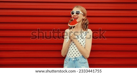 Summer portrait of happy smiling young woman with fresh juicy fruits, lollipop or ice cream shaped slice of watermelon wearing heart shaped sunglasses with cool girly hairstyle on red background