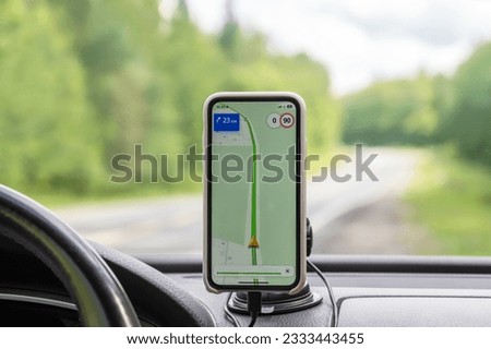 Smartphone in a holder with navigation app on screen on the dashboard of a car on the background of windshield with road landscape behind it.