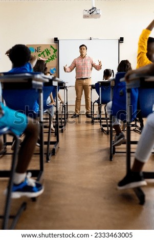 Diverse male teacher and elementary schoolchildren raising hands in class. School, learning, childhood and education, unaltered.
