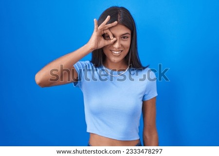 Brunette young woman standing over blue background doing ok gesture with hand smiling, eye looking through fingers with happy face. 