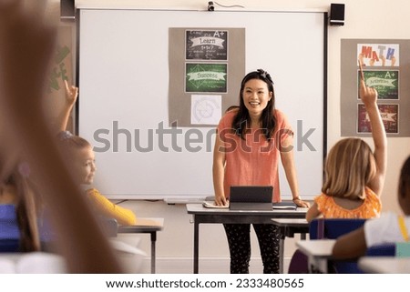 Diverse female teacher with tablet and elementary schoolchildren raising hands in class. School, learning, childhood, communication and education, unaltered.