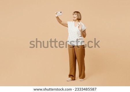 Full body blonde elderly woman 50s years old wear casual clothes doing selfie shot on mobile cell phone post photo on social network isolated on plain pastel light beige background. Lifestyle concept