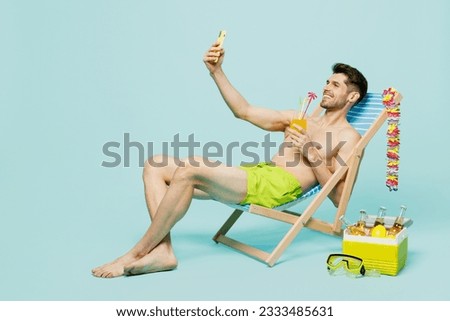 Full body young man wear green shorts swimsuit relax near hotel pool sit in deckchair do selfie shot mobile cell phone drink cocktail isolated on plain blue background. Summer sea rest sun tan concept