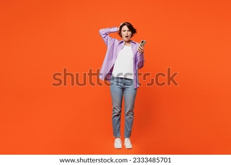 Full body angry mad astonished sad young woman she wear purple shirt white t-shirt casual clothes hold head use mobile cell phone isolated on plain orange background studio portrait. Lifestyle concept Royalty-Free Stock Photo #2333485701
