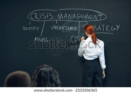 Young woman, employee writing on black board plan of crisis management. Meeting with colleagues for discussion and planning. Concept of business, planning, strategy, brainstorming, analytics, ad Royalty-Free Stock Photo #2333506365