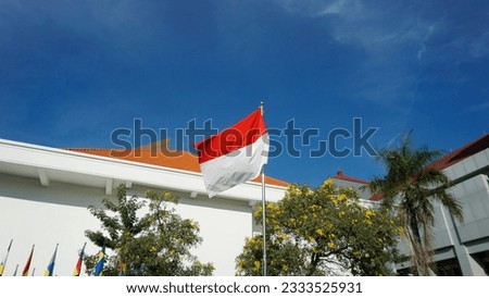 Indonesia flag fluttering on a pole in front of the building. Indonesian state flag, red and white. Blue and clear sky. 