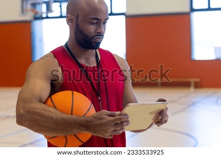 Focused biracial male coach holding basketball and using tablet at gym. Sport, activity, technology and lifestyle, unaltered.