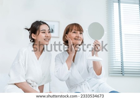 Asian woman looking at mirror after apply lotion on face with mother. Attractive young girl and senior older mom in bathrobe sit on bed, feel happy and proud of healthy beauty treatment and skin care.