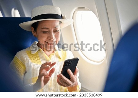 An attractive and happy senior Asian female passenger using her smartphone at her window seat on a flight, travelling for her summer trip. Royalty-Free Stock Photo #2333538499