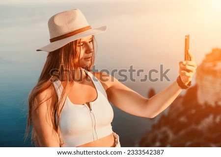 Selfie woman in a hat, white tank top, and shorts captures a selfie shot with her mobile phone against the backdrop of a serene beach and blue sea.