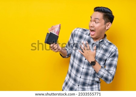 Surprised handsome young Asian man wearing a white checkered shirt holding wallet with cash money isolated over yellow background. People Lifestyle Concept Royalty-Free Stock Photo #2333555771