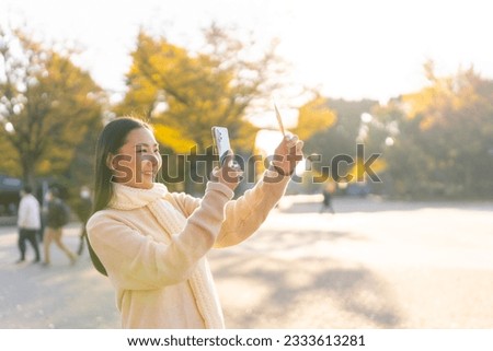 Asian woman using mobile phone taking picture of yellow leaf with sunlight during travel at public park in Tokyo city, Japan in autumn morning. Attractive enjoy outdoor lifestyle on holiday vacation.