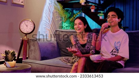 Happy Indian teenager couple sitting on cozy sofa watching favourite movie TV having fun enjoy laughing weekend together. Smilling teen boyfriend girl friend hold popcorn bowl eating snack at home