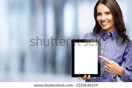 Happy smiling brunette businesswoman showing blank tablet pc monitor, with copy space area for some text, advertising or slogan, over blurred modern office interior background. Business woman indoors.