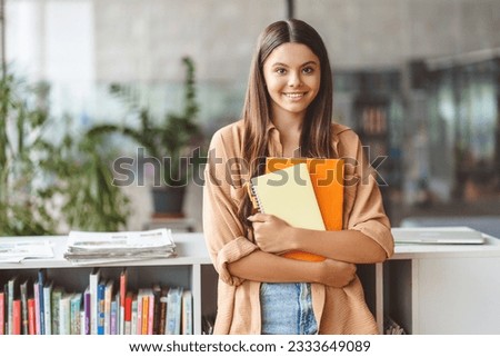 Portrait of smiling smart teenage girl holding book looking at camera standing in modern classroom, copy space. Back to school, education concept