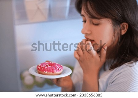 Young woman with tasty doughnut near open fridge in kitchen at night, closeup Royalty-Free Stock Photo #2333656885