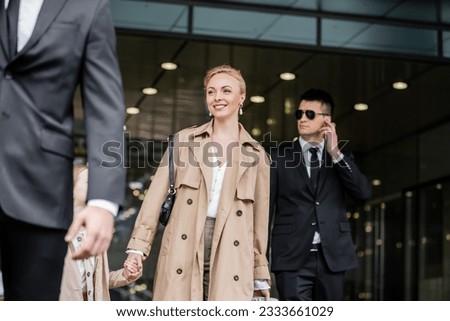 professional bodyguards protecting smiling blonde woman and preteen kid, successful mother and daughter in trench coats standing near hotel, safety and protection, private security, guards Royalty-Free Stock Photo #2333661029