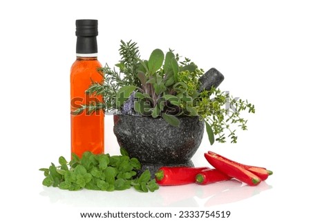 Chili oil in a bottle with fresh red pepper chillies and herb leaf sprigs of rosemary, sage, thyme in a granite mortar with pestle with loose oregano leaves, isolated over white background.