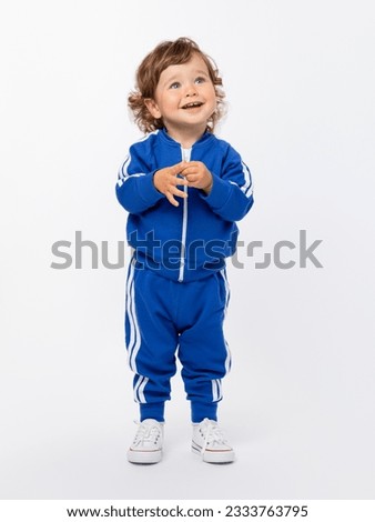 Childish naivety, purity. a curly-haired toddler boy of 1-2 years looks up expressively in a blue tracksuit and sneakers on a white background. Royalty-Free Stock Photo #2333763795