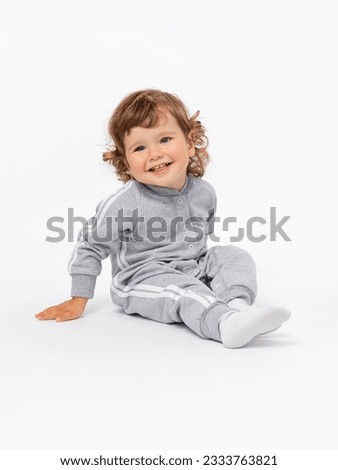 The toddler is 2 years old. A joyful boy is sitting on the floor and laughing with his head tilted with curly hair, plump cheeks in a gray jumpsuit and socks on a white background. Royalty-Free Stock Photo #2333763821