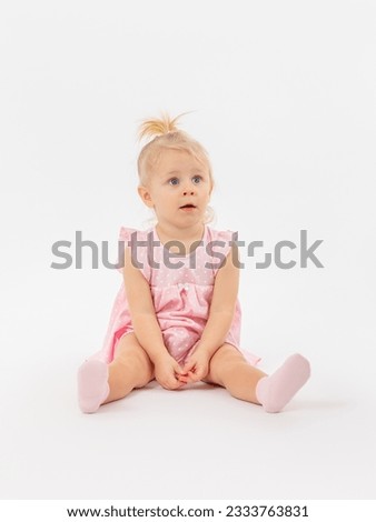 A little girl of 1-2 years old in a pink dress sits on the floor with her legs straight and looks in surprise at a white background. Copy space.