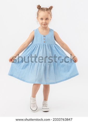 A 5-year-old girl with a fashionable hairstyle poses on a white background and shows her blue summer dress. Looks expressively.