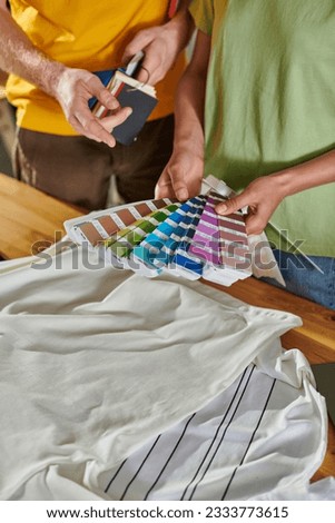 Cropped view of young african american craftswoman holding color swatches near colleague pointing with finger and standing near clothes in print studio, small business success concept