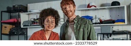 Positive and young multiethnic craftspeople looking at camera while standing together and working in blurred print studio, ambitious young entrepreneurs concept, banner