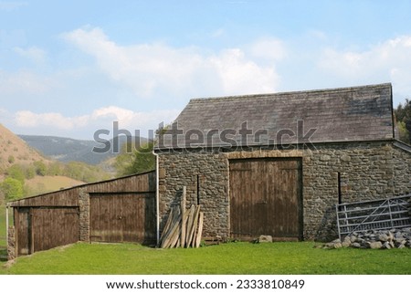 Old stone barn with oak doors and a slate roof with rural countryside to the rear against a pale blue sky.