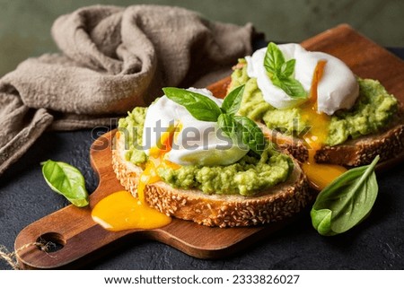 Smashed avocado sandwich with boiled poached egg with flowing  yolk and basil leaves. Dark green background. Royalty-Free Stock Photo #2333826027