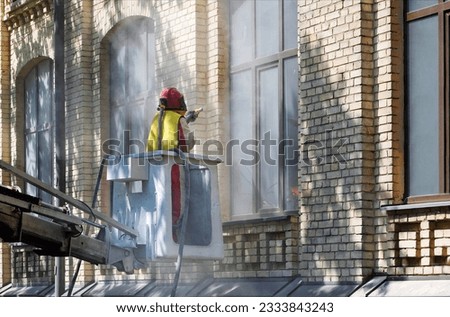 A worker in a protective suit, standing in a construction telescopic cradle, cleans the dirty coating from the brick wall of the facade of the house with a sandblaster. Royalty-Free Stock Photo #2333843243