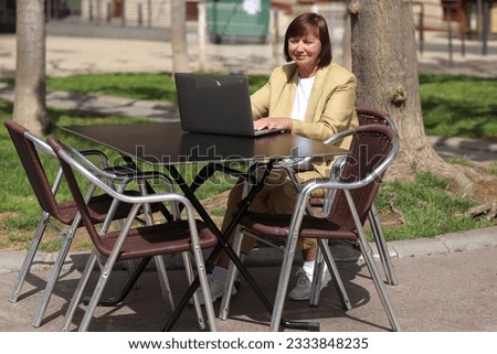 Elegant adult businesswoman works and type at laptop, making call on smartphone in an outdoor cafe at city. Concept of remote work communication from public place, digital freelance, modern lifestyle