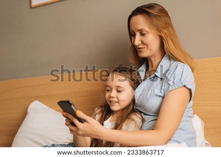 Happy young mother hugging to preschool daughter together browsing and surfing internet on mobile phone, chatting on video call sitting on bed in bedroom. Concept of family leisure activity at home.