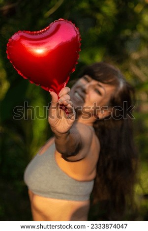 Blurred middle-aged brunette woman wearing a gray tank top, holding a metallic red heart balloon aloft in one hand, illuminated by sunlight.