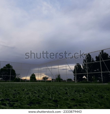 A picture of the dawn in a football field taken at ground level with a fence and trees at the background and a beautiful cloudy sky.