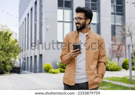 Successful young Hindu man walking outside office building, engineer software developer programmer smiling and happy using test app on phone, happy satisfied with result. Royalty-Free Stock Photo #2333879759
