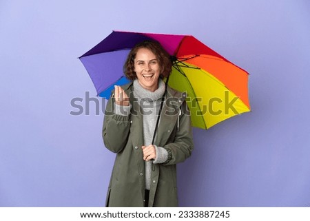 English woman holding an umbrella isolated on purple background making money gesture
