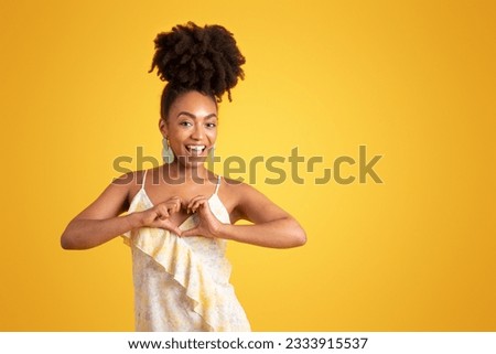 Happy young black lady in dress making heart sign with hands, isolated on yellow background, studio. Love, gratitude, recommendation, good news, romance emotions, ad and offer