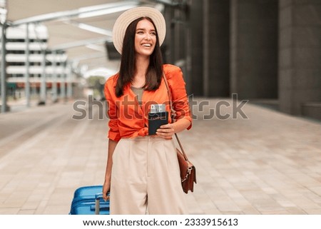 Happy Tourist Lady In Hat Holding Passport And Boarding Pass Walking With Luggage At Modern Airport Outdoors, Looking Away, Advertising Travel Offer. Passenger With Suitcase Arriving At Terminal Royalty-Free Stock Photo #2333915613